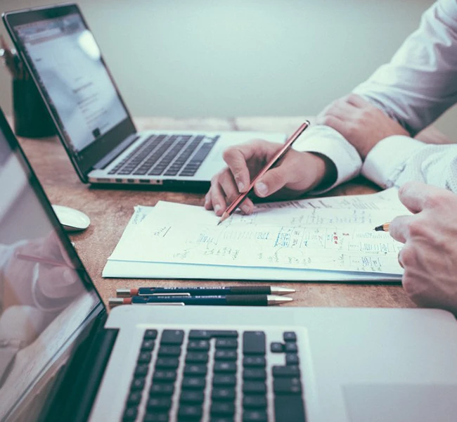 A hand holding a pen on top of papers between two laptop computers.